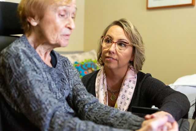 A woman with glasses and a pink scarf listens attentively to an older woman in a grey sweater while holding her hands.