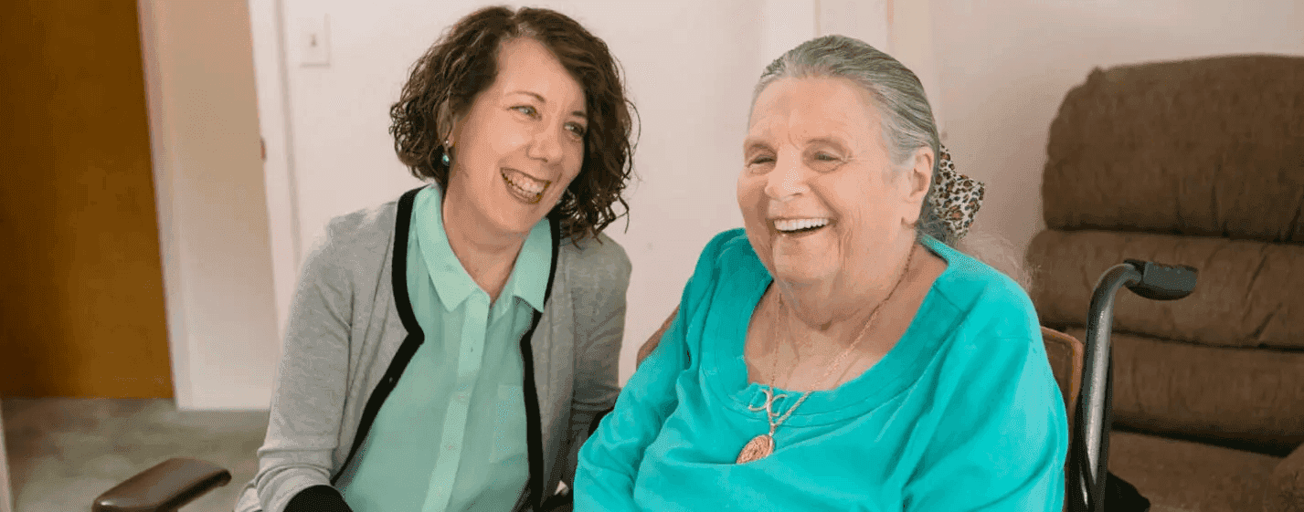 An older woman in a bright teal shirt and a younger woman with curly hair laugh together while sitting indoors.