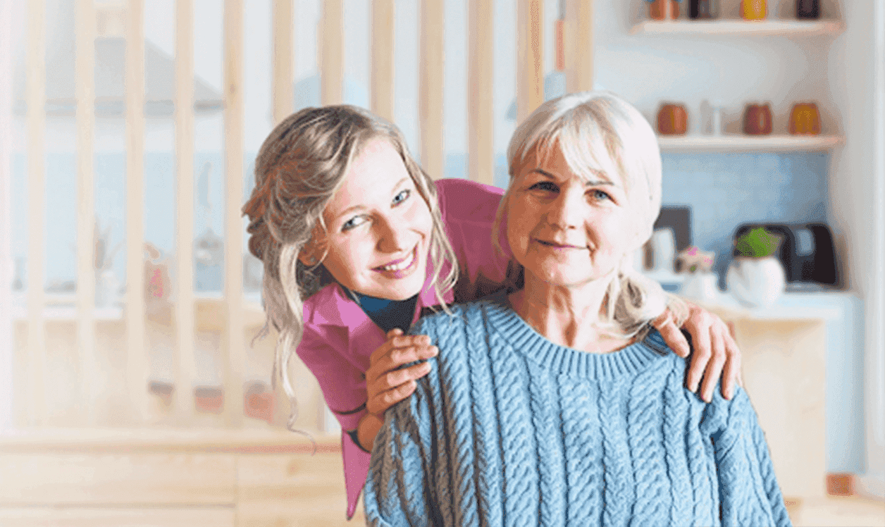 A smiling young female caregiver stands behind and gently places her hands on the shoulders of an elderly woman with short, light-blonde hair, in a warmly-lit home kitchen.