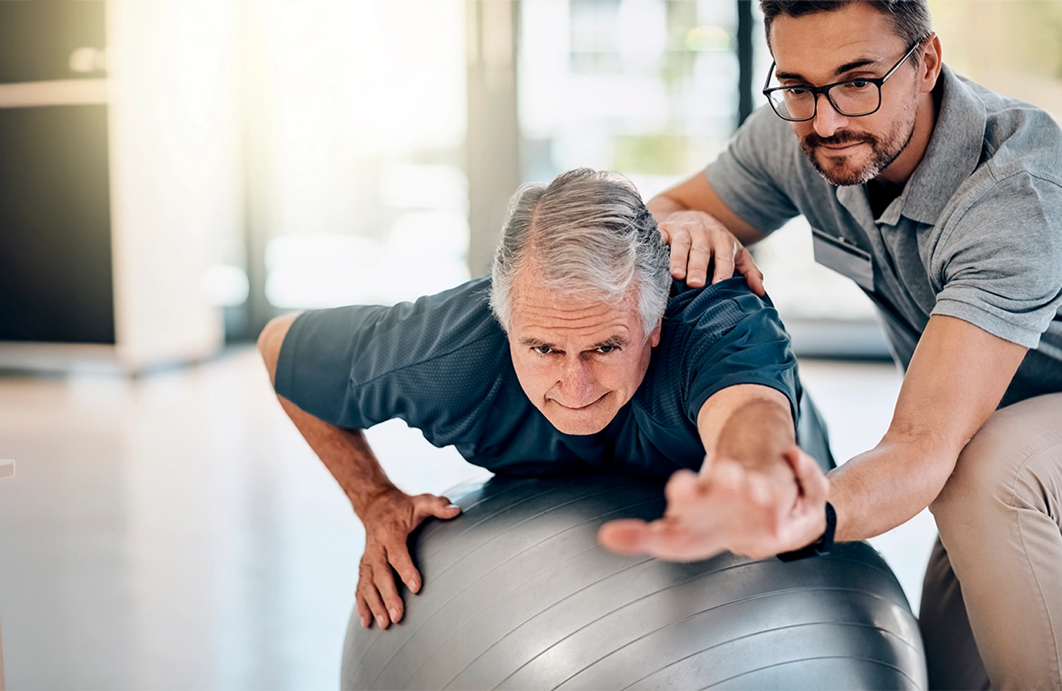 A senior man and his physical therapist doing balance exercises with a large stability ball in a sunlit living room.