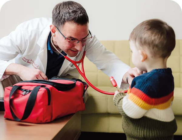 Male doctor using a stethoscope to examine a young child on a sofa during an in-home visit.