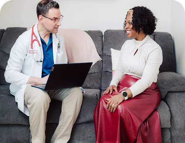 Male healthcare provider reviewing a laptop with a female patient on a sofa in her home.