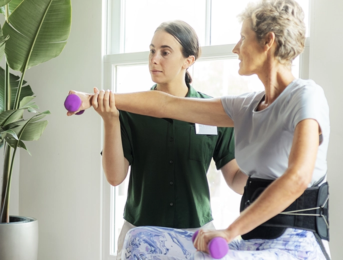 A female physical therapist guiding a mature woman through light arm exercises with small weights in a home setting.